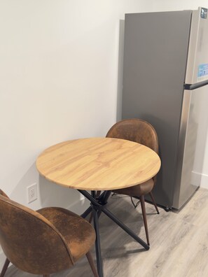 Uma pequena área de refeições com uma mesa redonda de madeira e duas cadeiras, uma geladeira e uma parede branca.