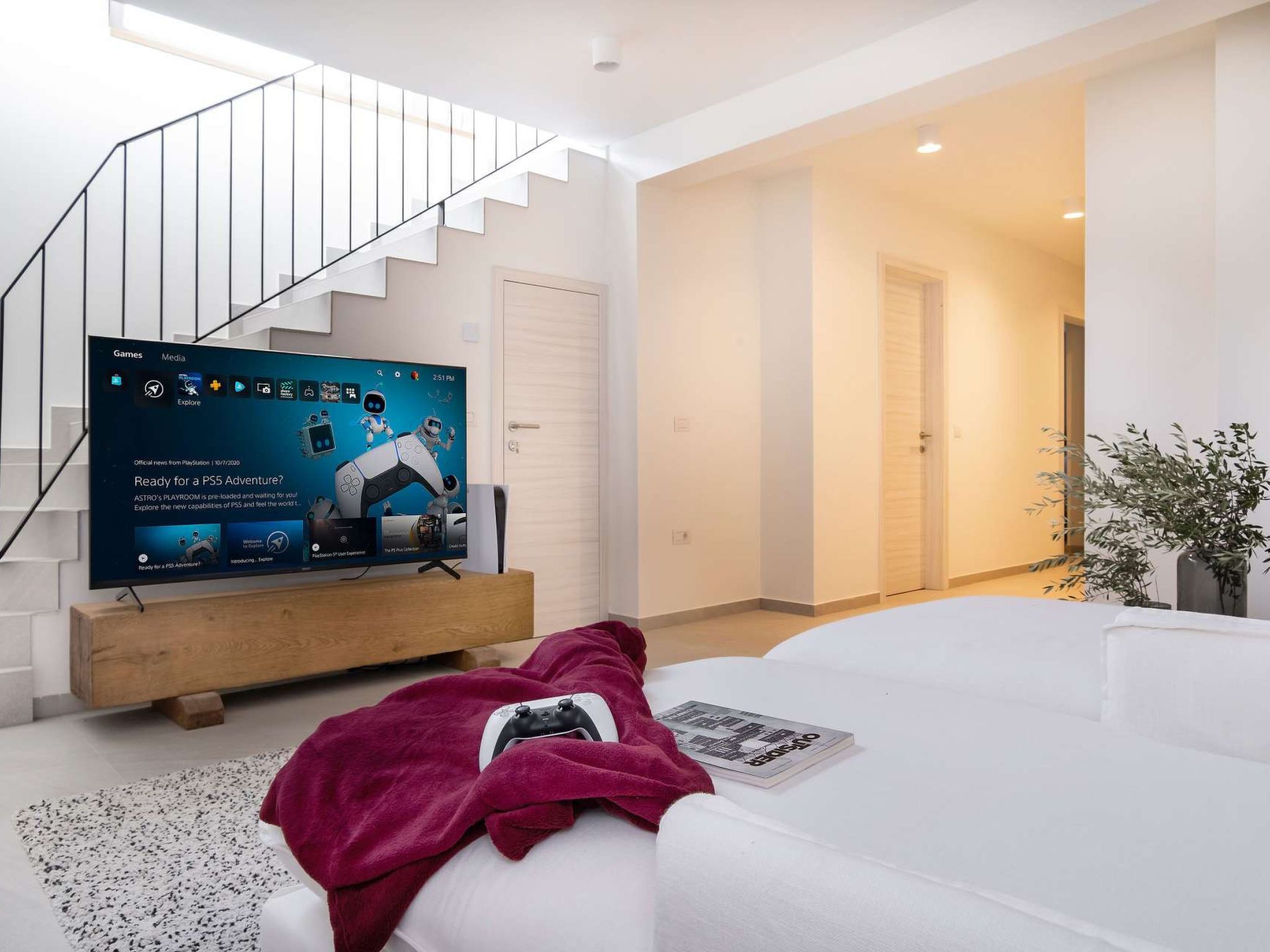 Ein modernes Wohnzimmer mit einem Flachbildfernseher auf einem Holzständer, einem weißen Sofa, einer Treppe und einer Tür zu einem anderen Raum.