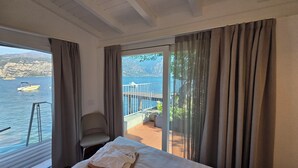 Habitación de hotel con cama, una silla, un balcón con vistas al mar y las montañas, y un ventanal con vista al agua.