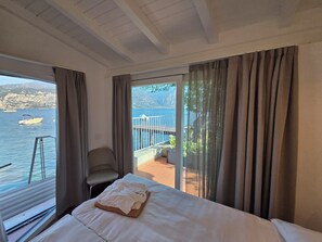 Habitación de hotel con cama, una silla, un balcón con vistas al mar y las montañas, y un ventanal con vista al agua.