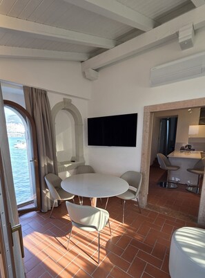 Una habitación con una mesa redonda, sillas, un televisor de pantalla plana, un ventanal con cortinas y vista al mar.