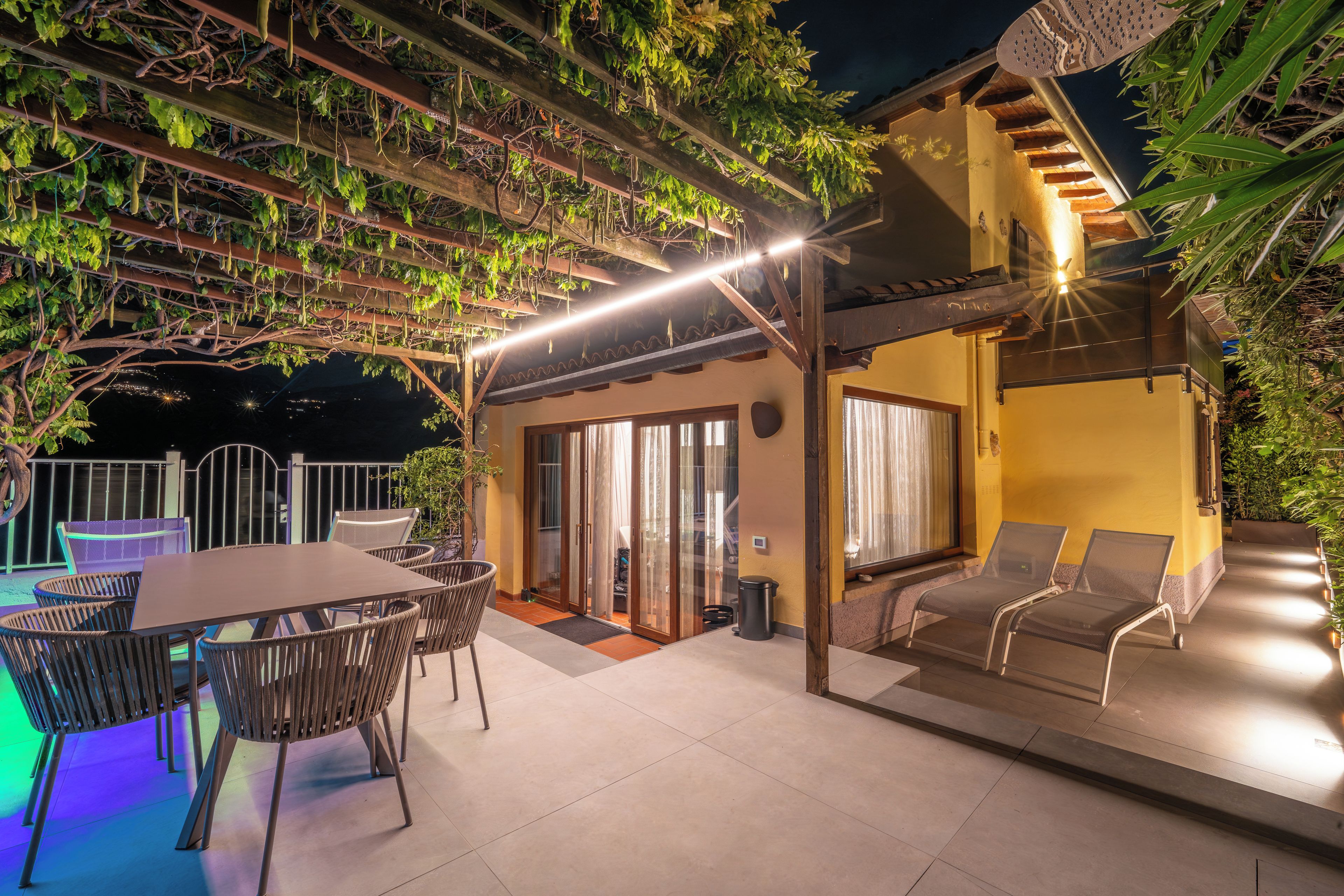 An outdoor patio with a dining table and chairs, a covered seating area with lounge chairs, and a pergola with climbing plants.