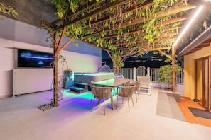 Een terras met een eettafel en stoelen, een ingebouwde barbecue en een pergola met klimplanten.
