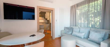 A modern living room with a white sofa, a dining table, and a TV mounted on the wall.