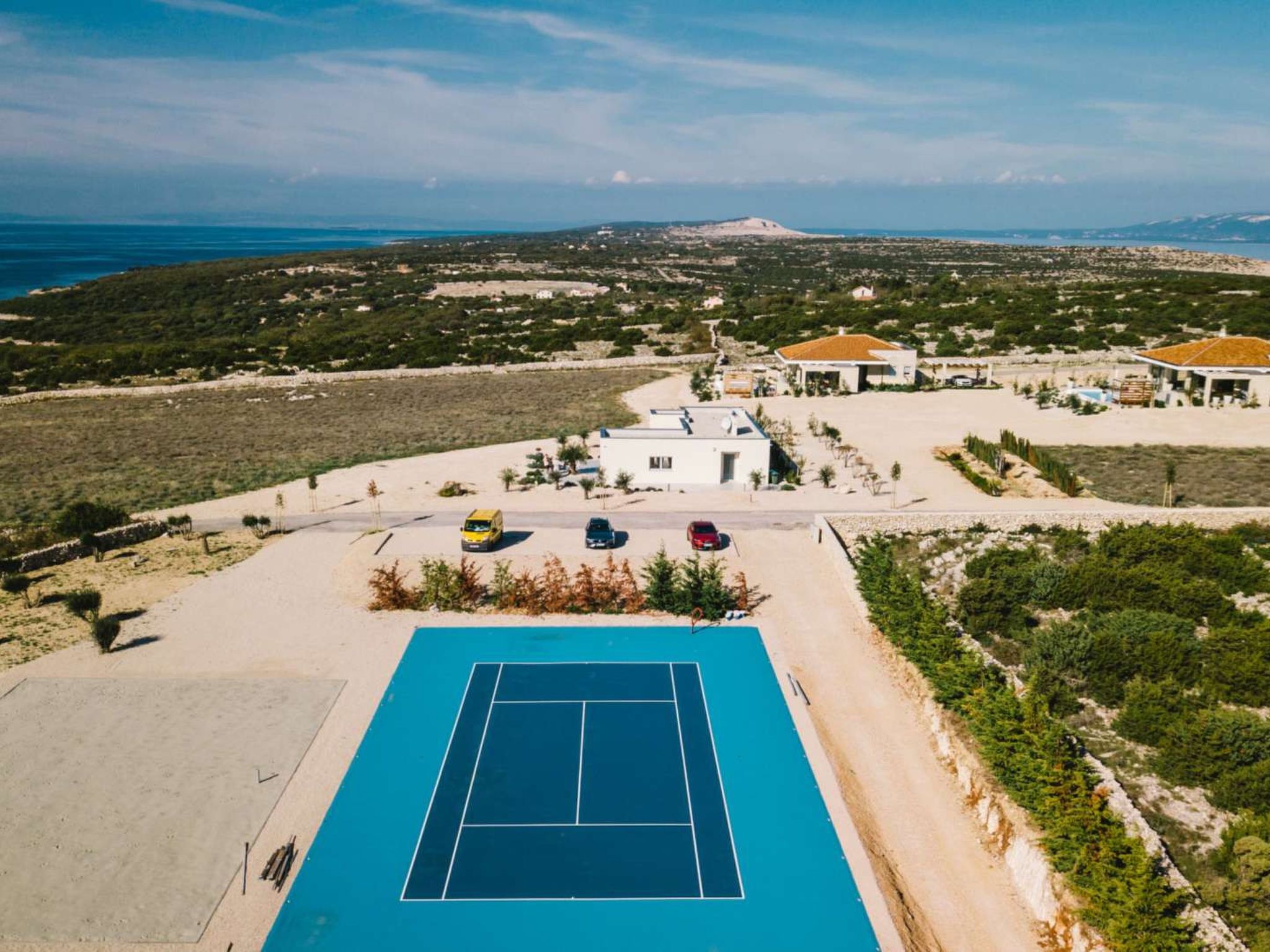 A tennis court with a clear view of the surrounding landscape, including a beach and distant hills.