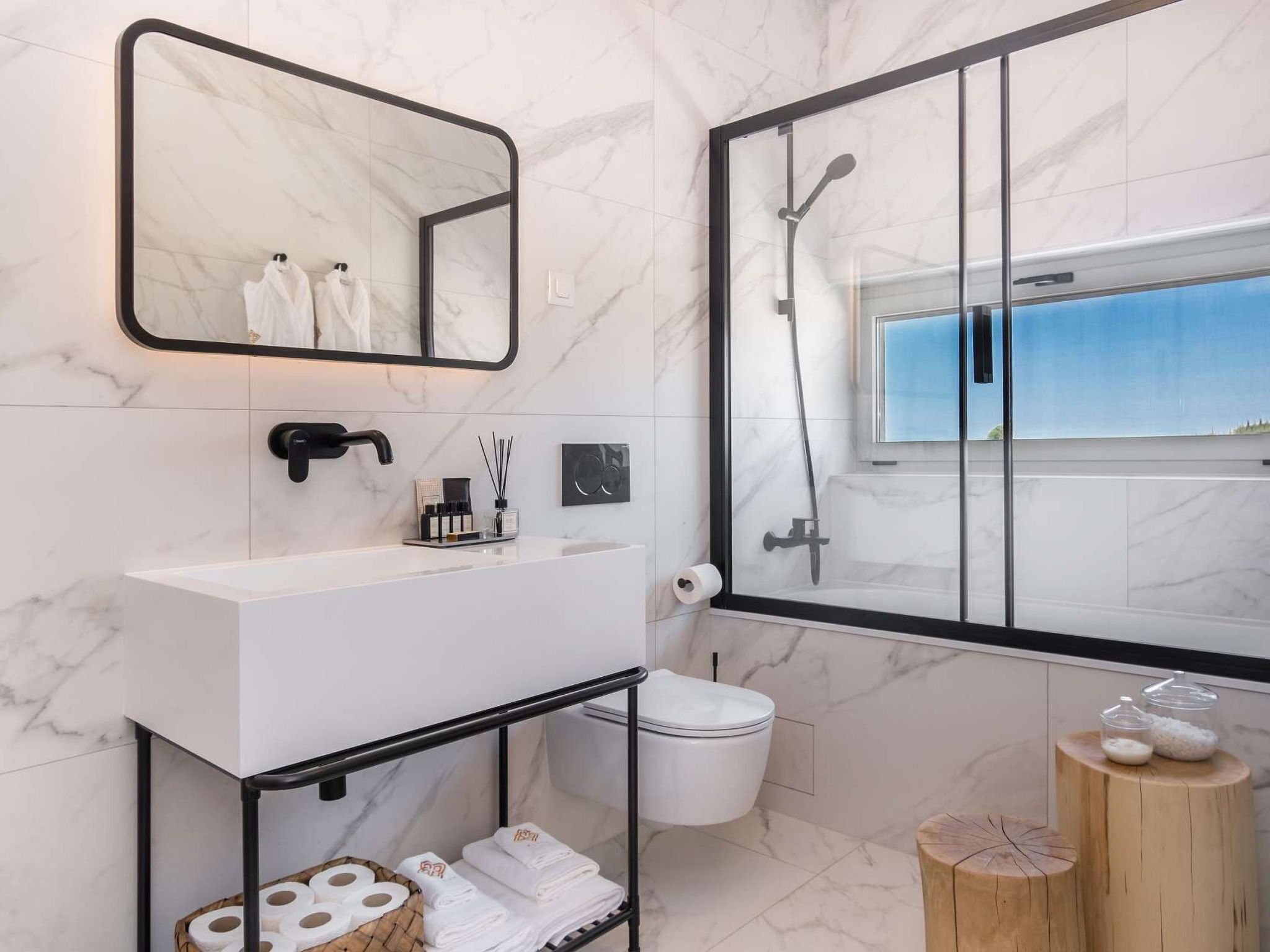 Une salle de bain moderne avec un sol en marbre, un lavabo blanc, un miroir à cadre noir, une douche en verre et un tabouret en bois sur lequel sont posés des articles de toilette.