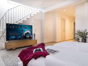 Ein modernes Wohnzimmer mit einem Flachbildfernseher auf einem Holzständer, einem weißen Sofa, einer Treppe und einer Tür zu einem anderen Raum.