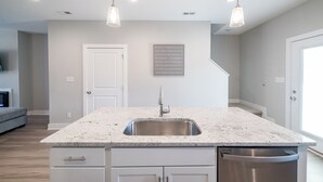 Una cocina con una isla central, pileta de acero inoxidable y lavavajillas. Hay lámparas colgantes sobre la isla y un cartel en la pared con mensajes positivos.