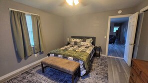 Un dormitorio con cama, banca, mesita de noche y una puerta que da a otra habitación.