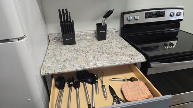 A kitchen with a granite countertop, a stove, and a drawer with utensils.