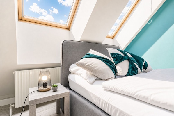 Un dormitorio con tragaluz, una mesita de noche con lámpara y una cama con almohadas.