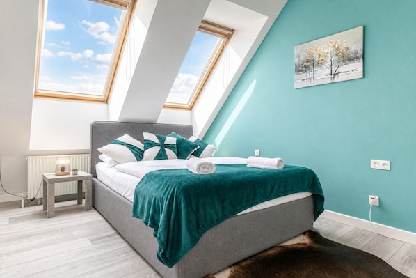 A bedroom with a skylight, a bed with a green blanket, a small bedside table, and a painting on the wall.