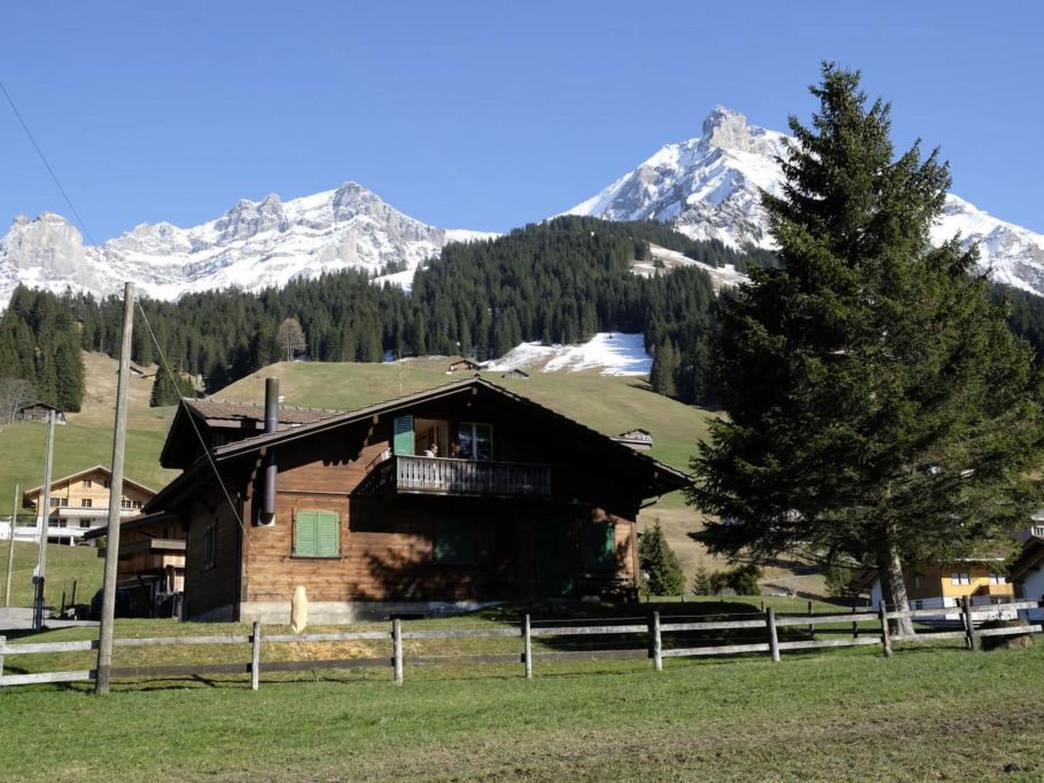 A wooden cabin with a balcony, surrounded by a fenced yard, set against a backdrop of snow-capped mountains and a clear sky.