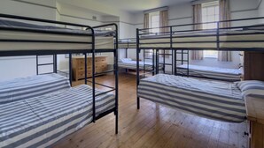 A dormitory room with bunk beds, wooden floors, a dresser, and a window with curtains.