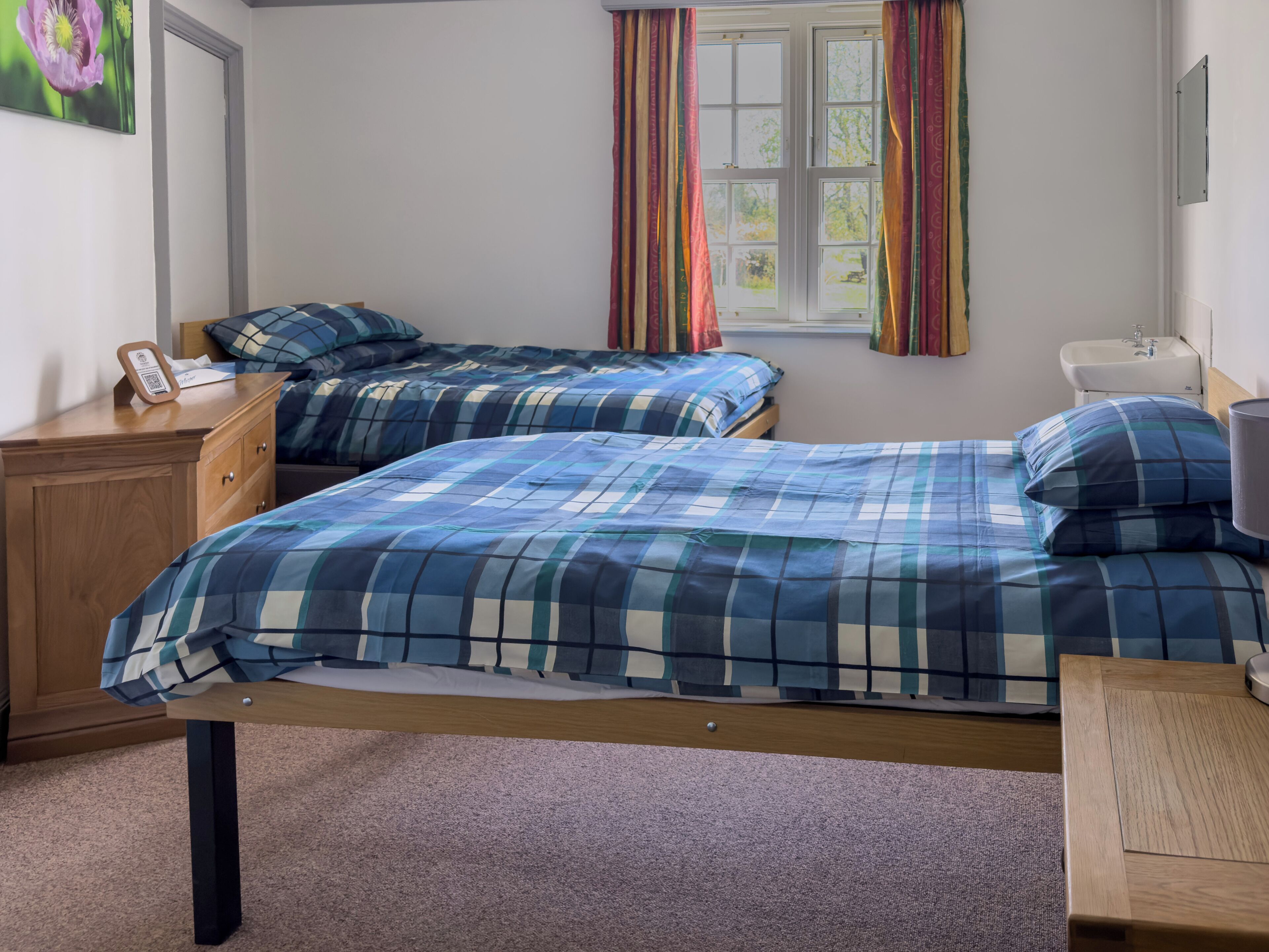 A bedroom with two beds, a wooden dresser, a bedside table, a window with curtains, and a sink.
