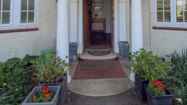 A building entrance with a classic architectural style, featuring a central doorway flanked by columns, a tiled entrance, and potted plants.