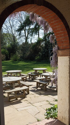 Un patio al aire libre con mesas y bancos de madera, rodeado por un arco de ladrillo.