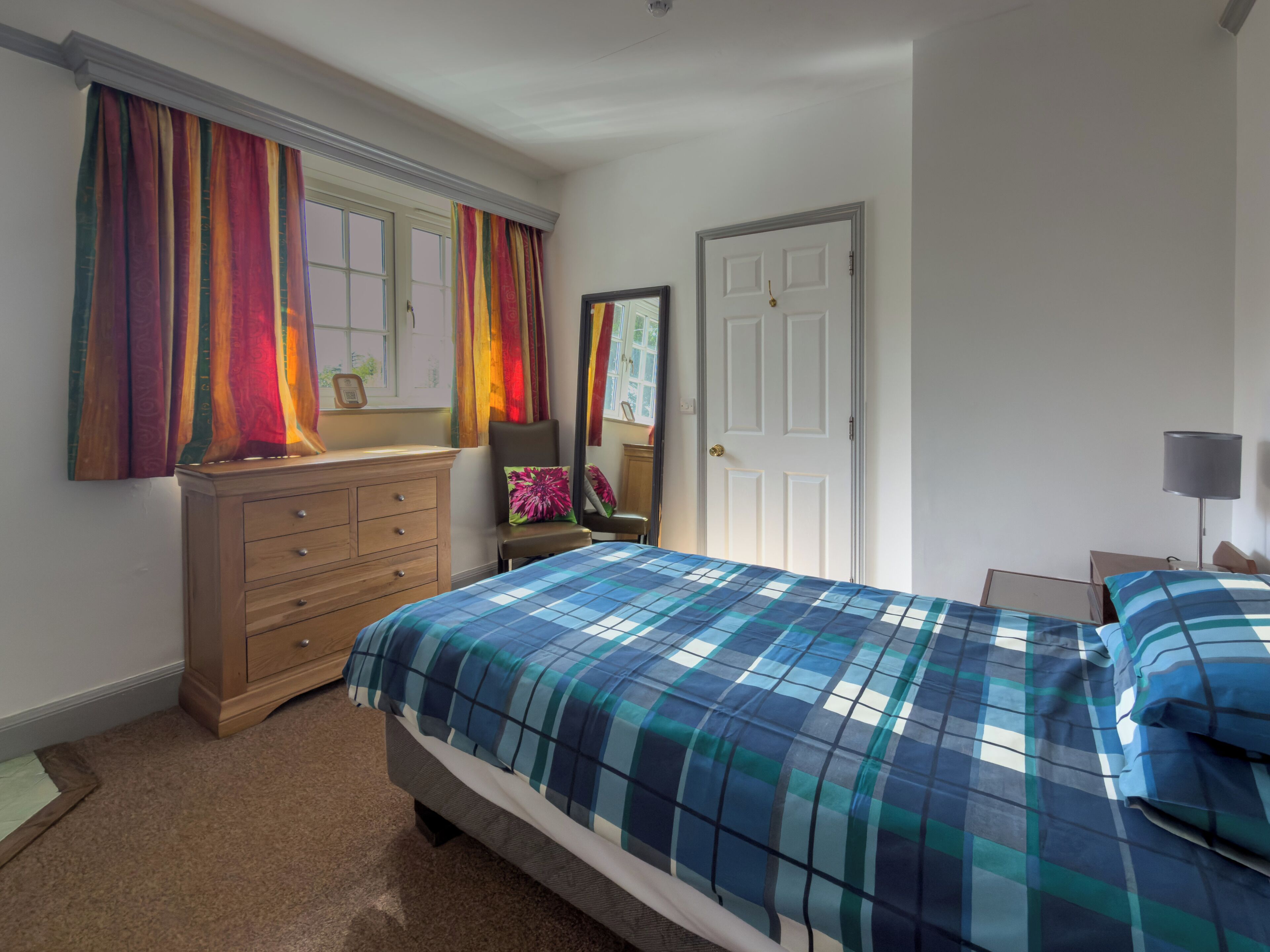 Un dormitorio con cama, cómoda, ventana con cortinas, puerta y mesita de noche con lámpara.