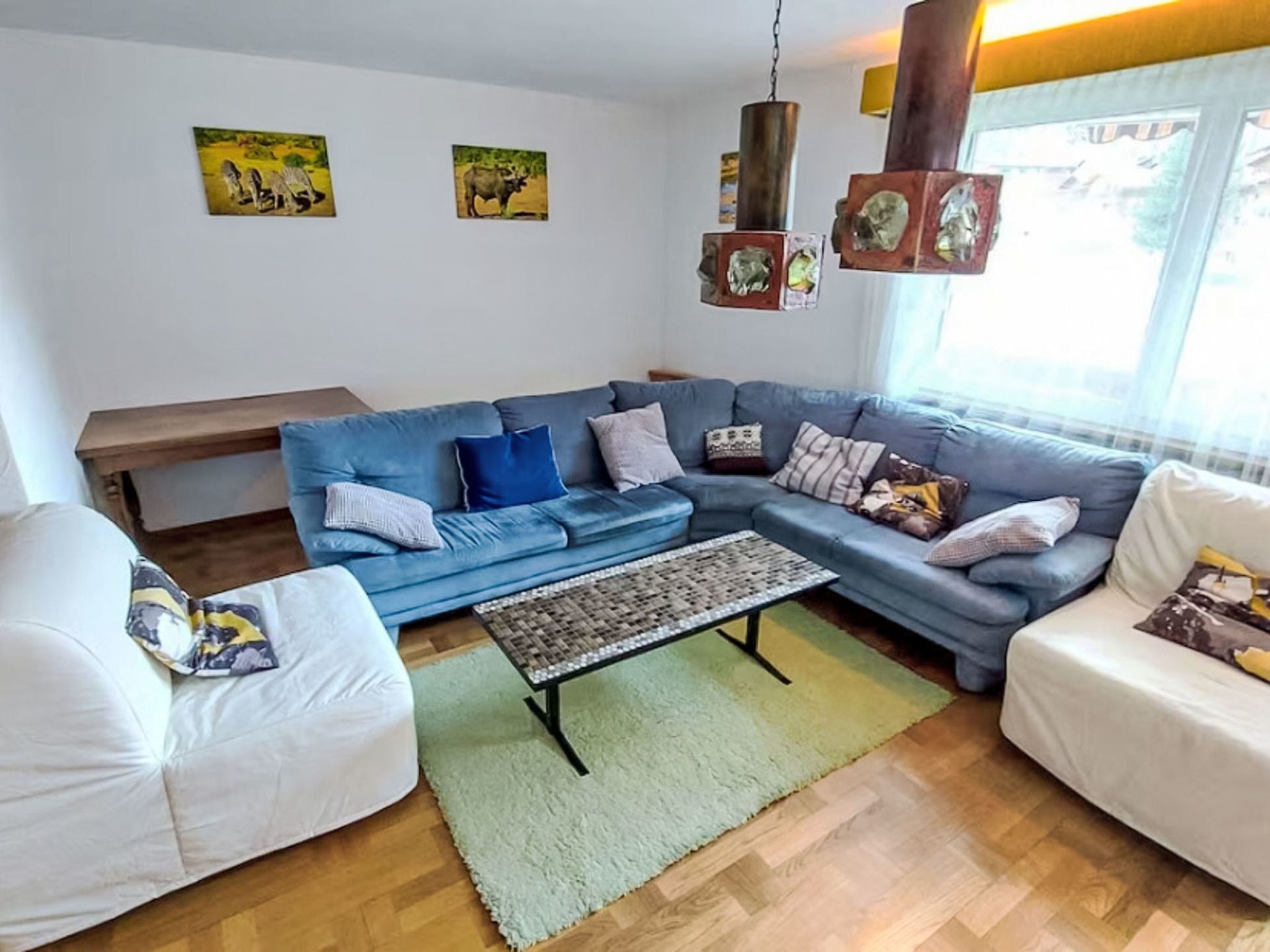Un salon avec un canapé d’angle bleu, un fauteuil blanc, une table basse en osier et un tapis vert. Des tableaux ornent le mur, et un luminaire suspendu éclaire la pièce.