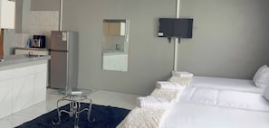 A hotel room with a kitchenette, a bed with white bedding, a small table, and a flat-screen TV mounted on the wall.