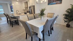 Uma sala de jantar com uma mesa posta para seis pessoas, rodeada por cadeiras, em uma cozinha moderna com armários brancos e uma geladeira de aço inoxidável.