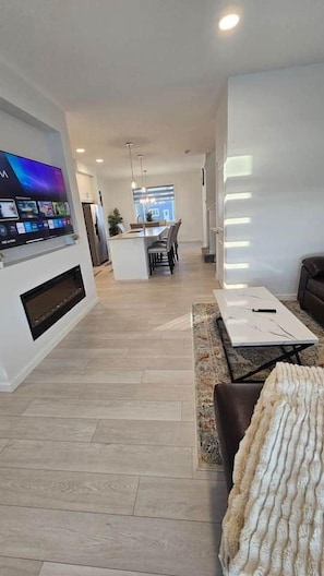 A modern living room with a flat-screen TV, a dining area with a table and chairs, and a kitchen in the background.