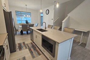A modern kitchen with a central island, a dining area with chairs, a staircase, and a clock on the wall.