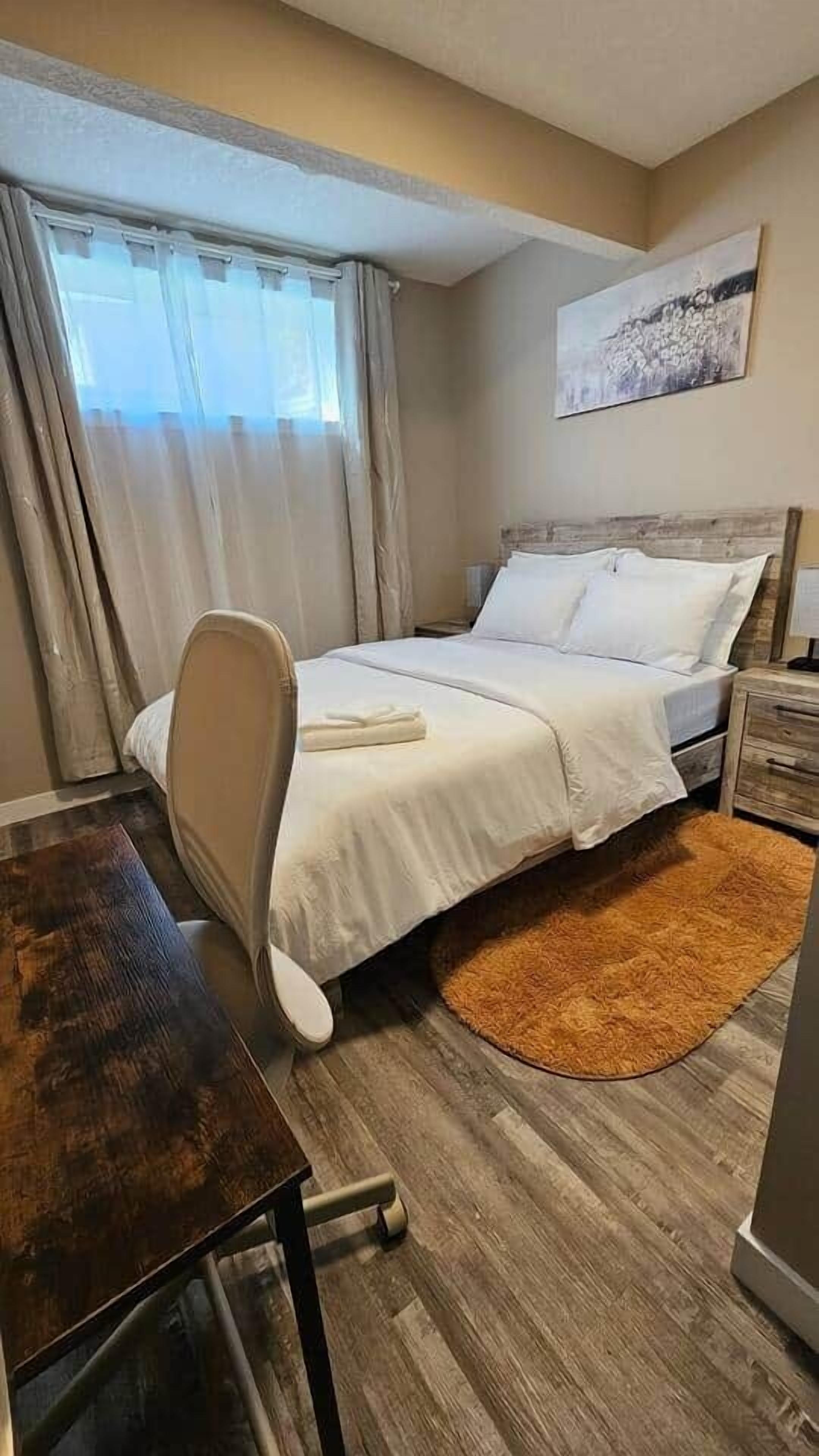 A hotel room with a bed, a desk, a chair, a wooden floor, a window with curtains, and a painting on the wall.
