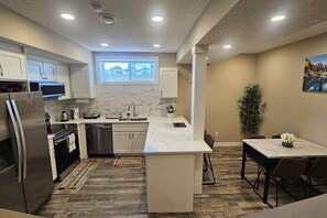 A compact kitchen with a built-in oven, a dishwasher, and a refrigerator. There's a small window above the sink, and a dining area with a table and chairs. - Luxury suite with 2-bedroom and 2 beds + Free Wifi (Calgary)