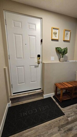 A white door with a doorknob, a black doormat with 'WELCOME' text, a wooden bench, and two framed botanical prints on the wall. - Luxury suite with 2-bedroom and 2 beds + Free Wifi (Calgary)
