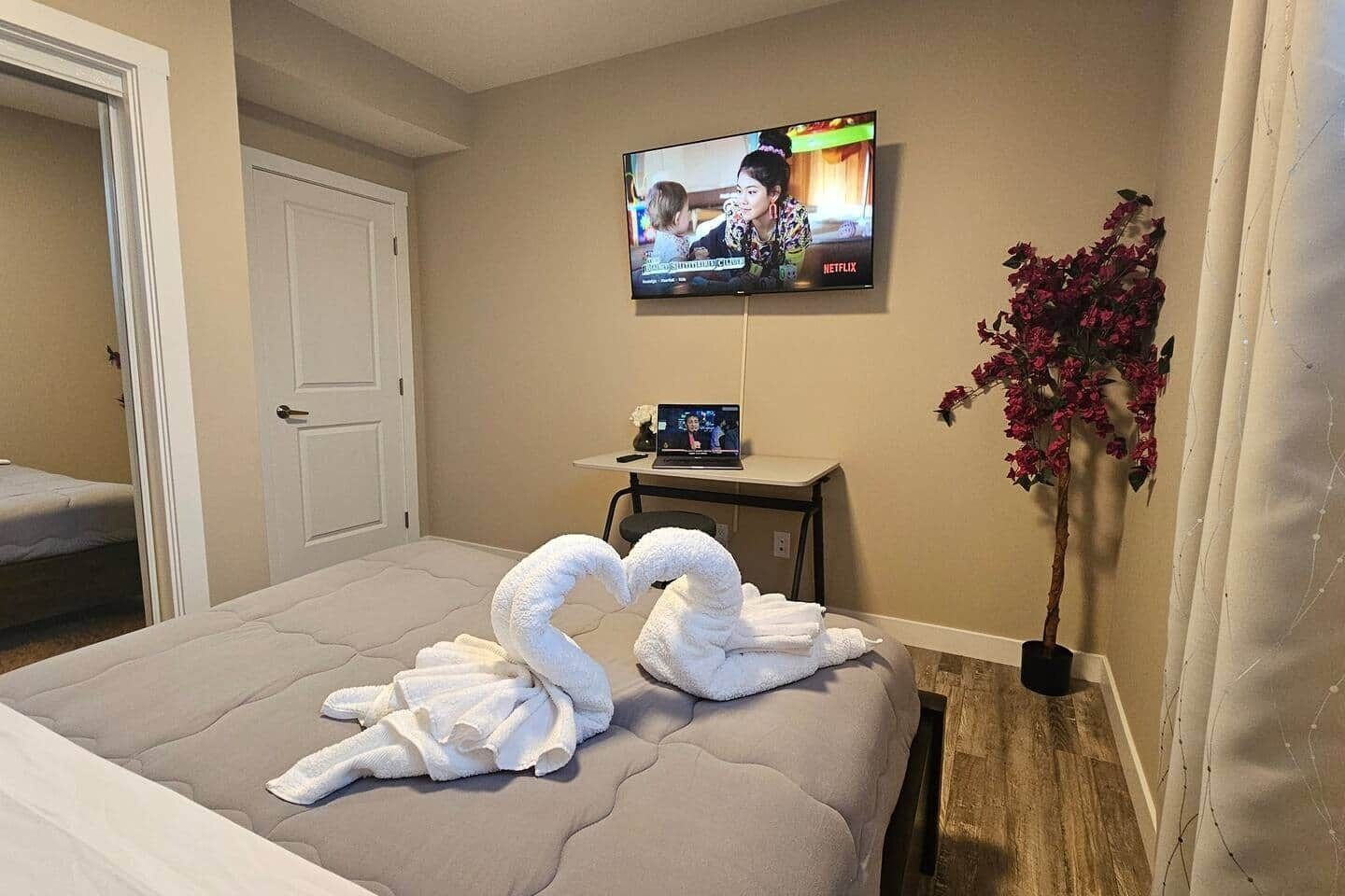 Une chambre d’hôtel avec un lit, un téléviseur fixé au mur, une petite table avec un ordinateur portable et un vase rempli de fleurs.