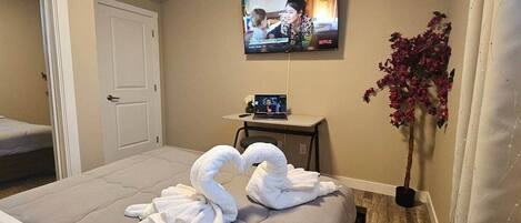 Une chambre d’hôtel avec un lit, un téléviseur fixé au mur, une petite table avec un ordinateur portable et un vase rempli de fleurs.