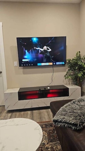 A flat-screen TV mounted on a wall, a leather ottoman, a round white table, and a potted plant. - Luxury suite with 2-bedroom and 2 beds + Free Wifi (Calgary)