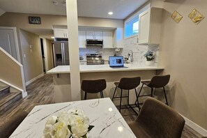 A modern kitchen with white cabinets, a stainless steel refrigerator, a microwave, and a laptop on the counter. There are four bar stools and a table with a vase of flowers. - Luxury suite with 2-bedroom and 2 beds + Free Wifi (Calgary)