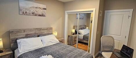 A bedroom with a wooden headboard, a bed with white linens, a desk with a laptop, and a mirror reflecting the room.