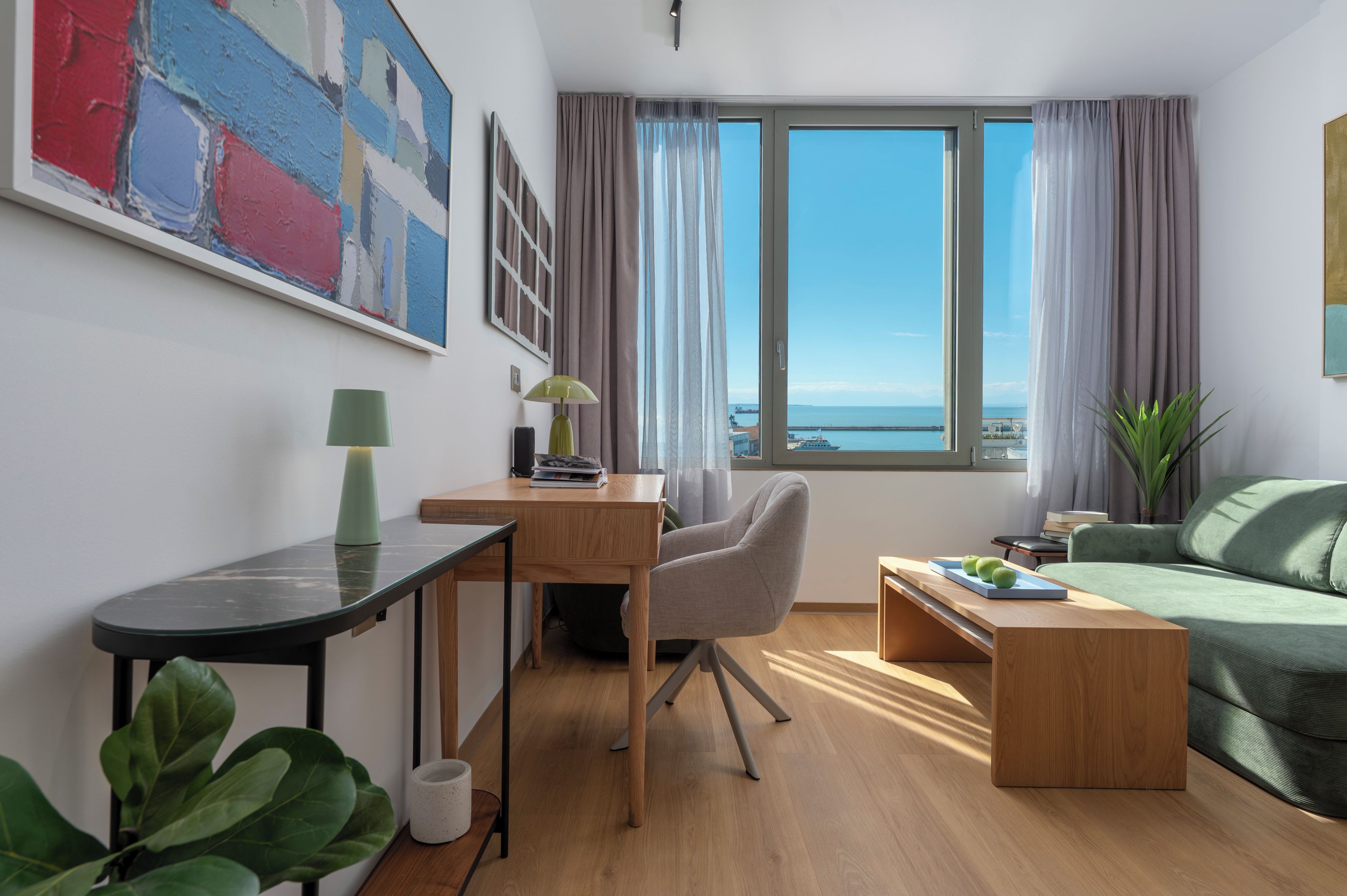 A modern living room with a desk, a green sofa, a view of the sea, and a painting on the wall.