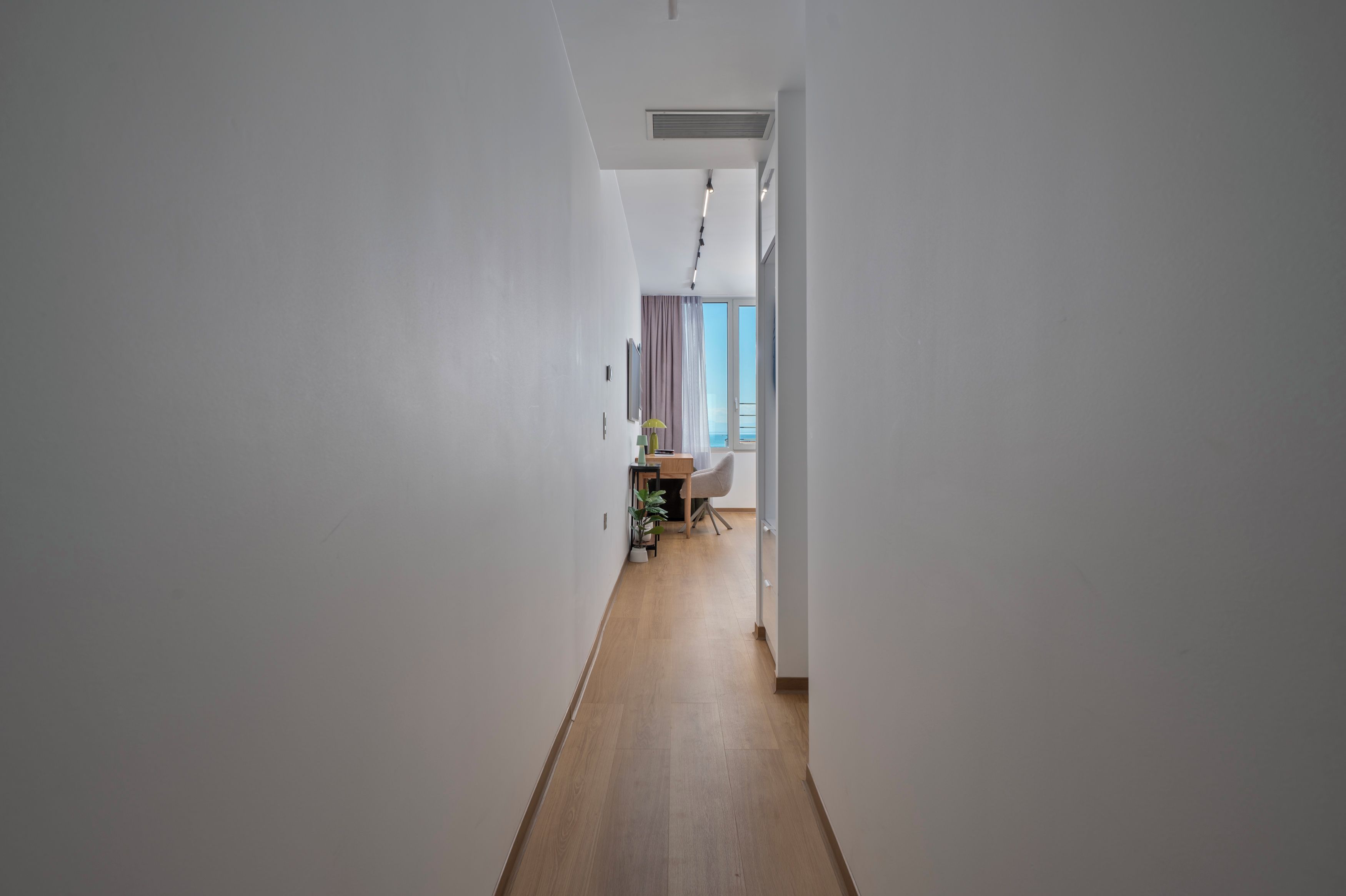 A hallway with a wooden floor leading to a room with a desk and chair, a plant, and a view through a window.