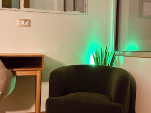 A room with a dark green upholstered chair, a wooden side table, and a green ambient light source near a window. - Blue Elegant Suite (Thessaloniki)
