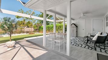 A covered patio area with a dining table and chairs, a seating area with a patterned rug, and a view of a garden and house in the background.