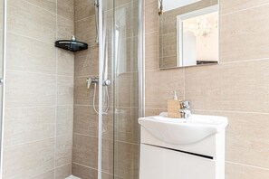 A modern bathroom with a glass shower enclosure, a white sink with a wooden soap dispenser, and a mirror reflecting a chandelier.