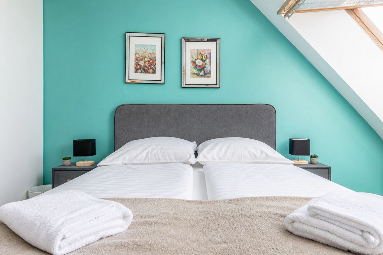 Ein Bett mit weißen Leinentüchern, zwei Nachttischlampen und eine türkisfarbene Wand mit zwei gerahmten Bildern.