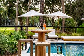 Ein Poolbereich mit Holztisch, zwei Stühlen, einer Weinflasche und zwei Sonnenschirmen.