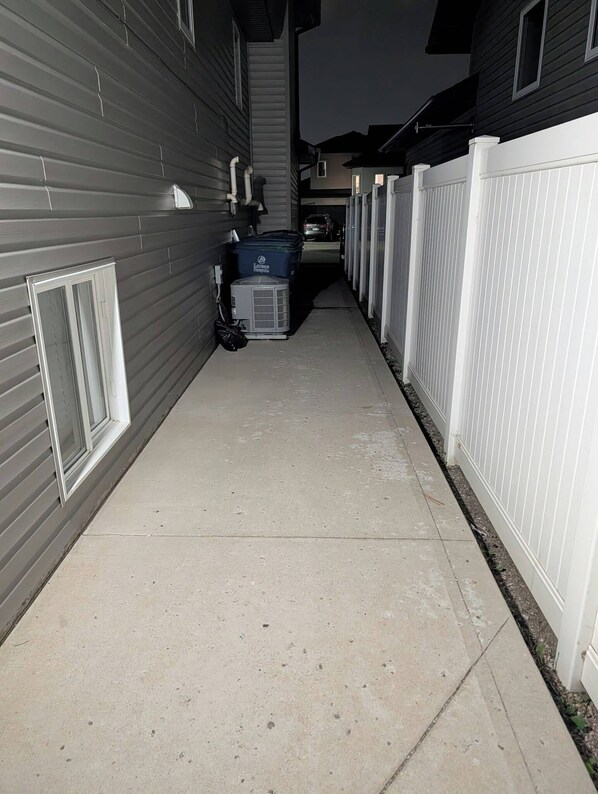 A narrow concrete pathway flanked by buildings with siding and a white fence.