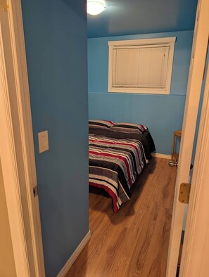 A narrow hallway leading to a bedroom with a bed, a window with blinds, and a wooden floor.