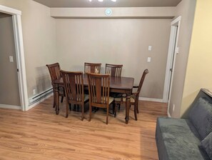 A dining area with a wooden table and chairs, a gray couch, and a door on the right. - Modern two bedroom legal suite in beautiful evergreen, Saskatoon (Saskatoon)