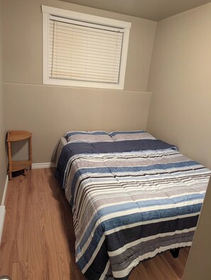 A small, single-bed room with a wooden floor, a window with blinds, and a small wooden chair.