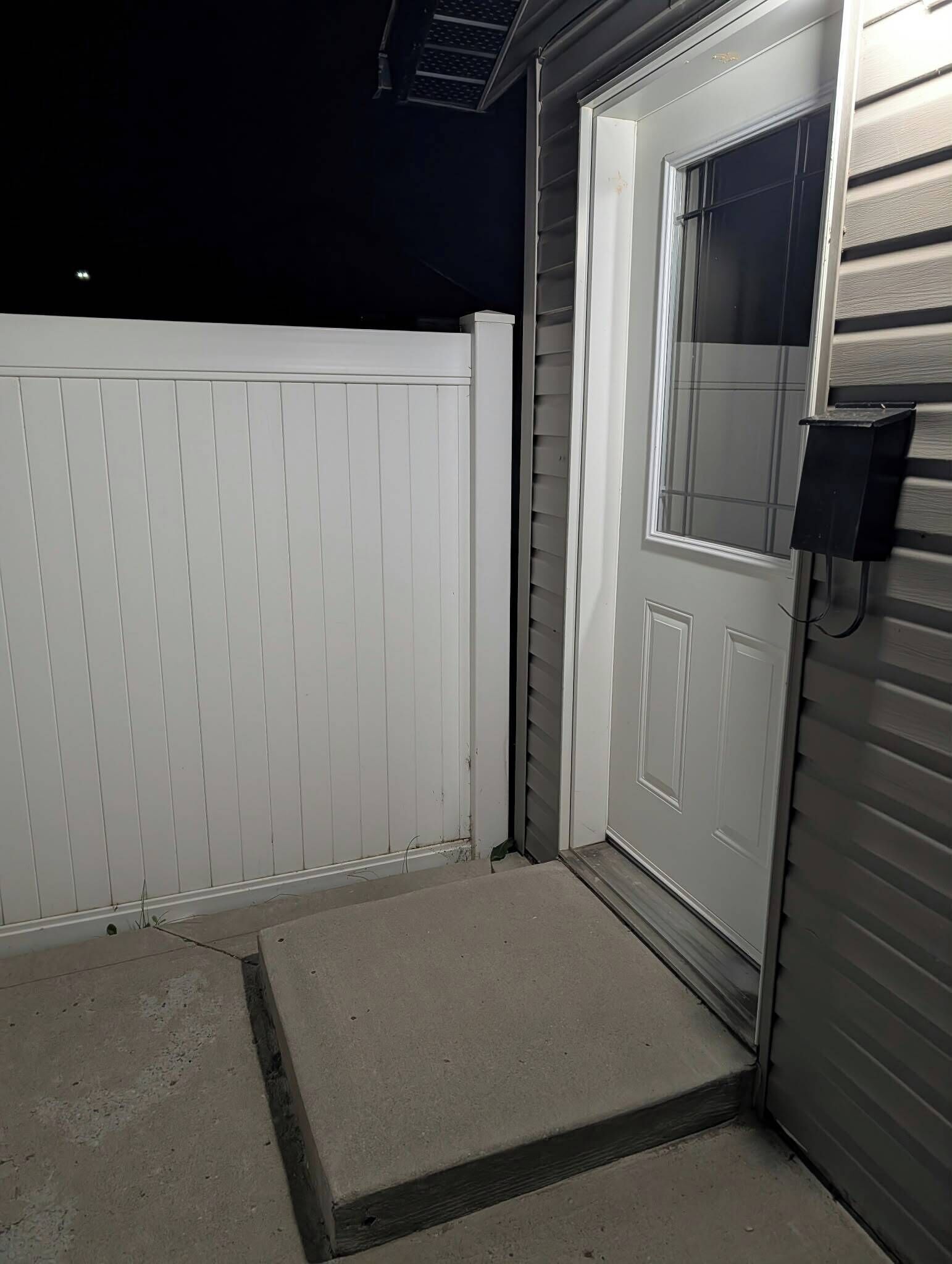 Ett garage med en vit dörr, ett betongsteg som leder upp till dörren och en metallanordning för att öppna garagedörren, placerad på höger sida.
