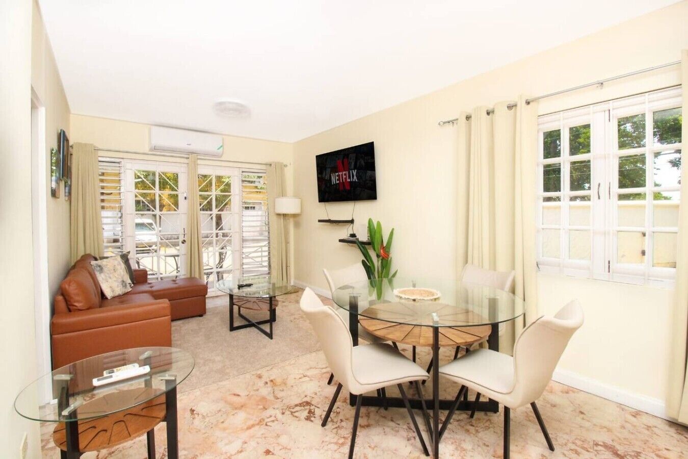 A modern living room with a glass dining table, white chairs, a brown leather sofa, and a flat-screen TV mounted on the wall.