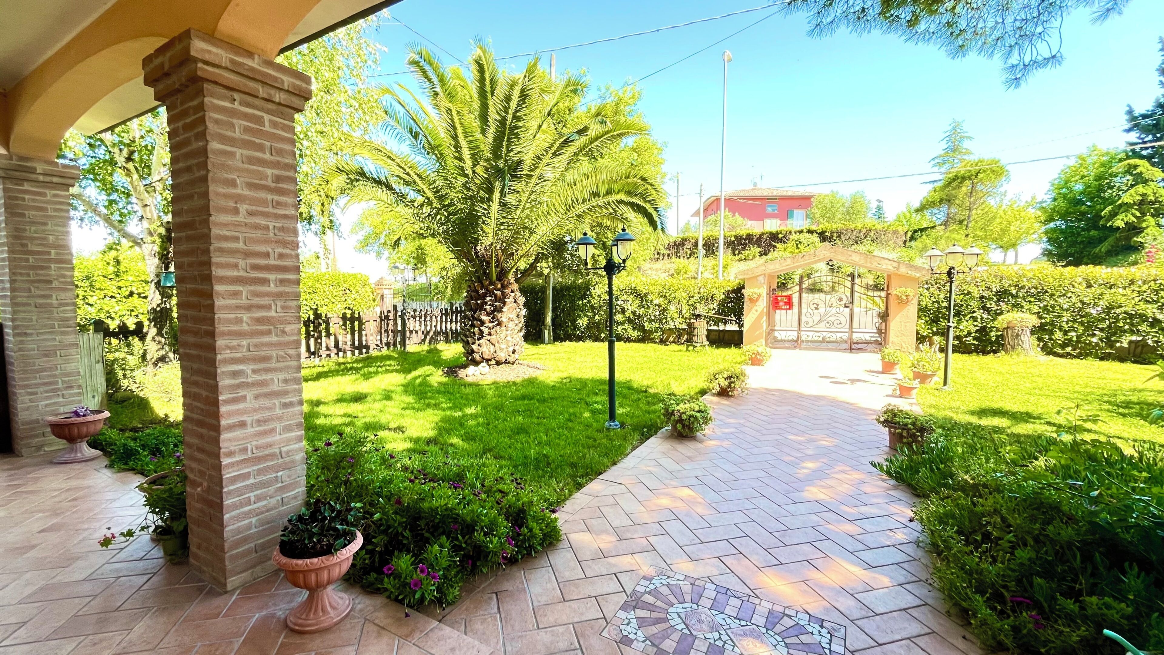 Un sendero en un jardín conduce a una entrada con portón, rodeado de setos cuidados y plantas en macetas.