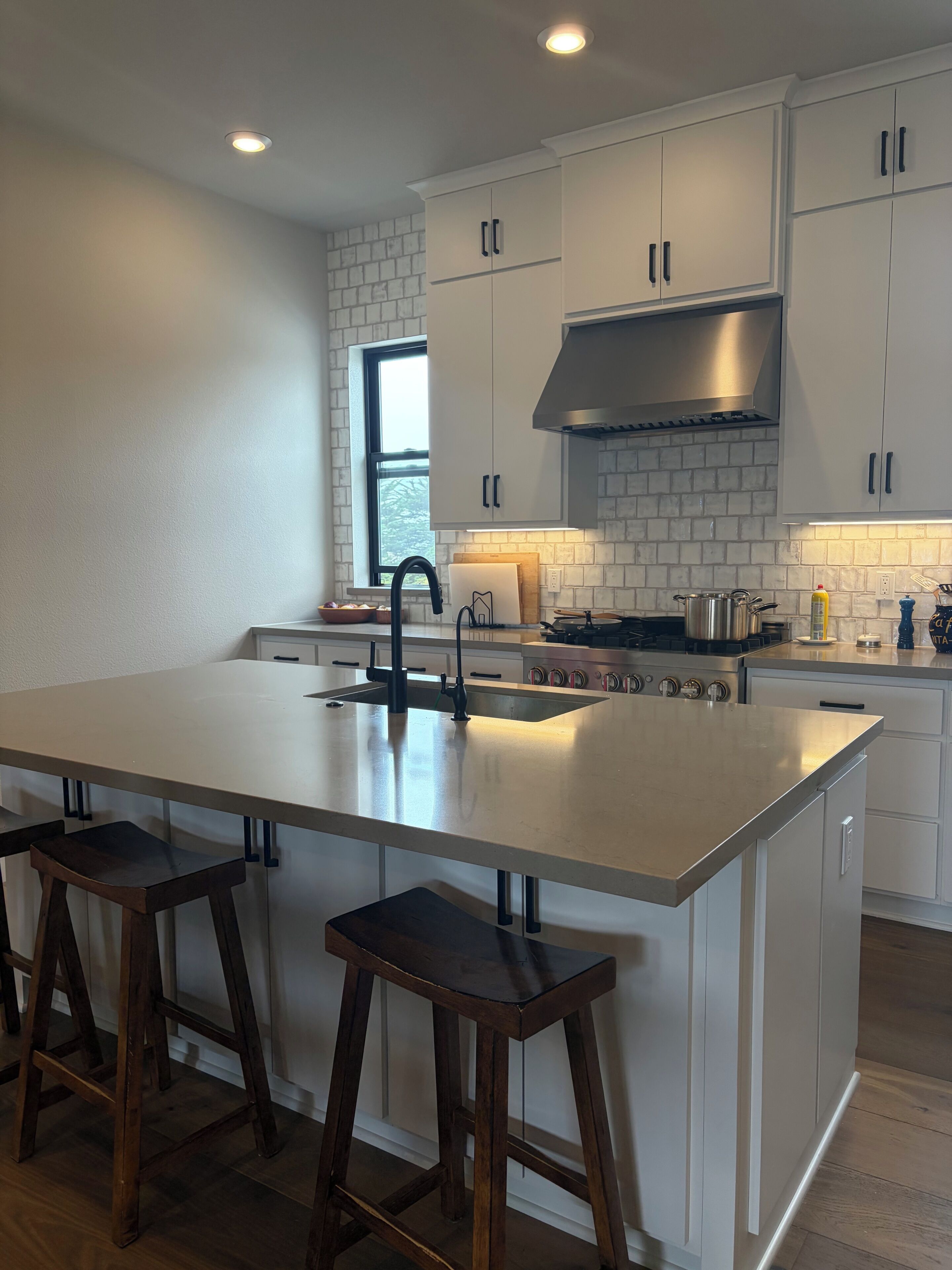 A modern kitchen with white cabinetry, a large island with bar stools, a stainless steel range hood, and a window providing natural light.